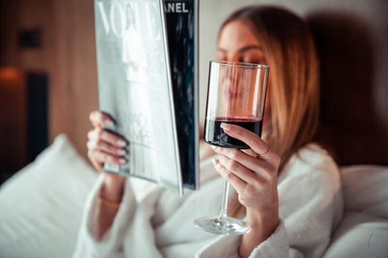 Woman laying on a bed in a bathrobe holding a glass of red wine and reading a magazine