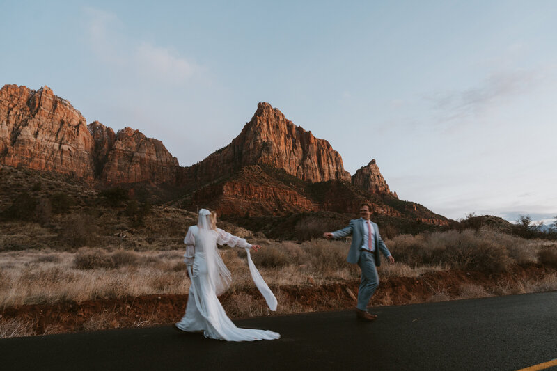 A couple eloping in their wedding attire running with mountains at sunset in the background at Zion Utah  