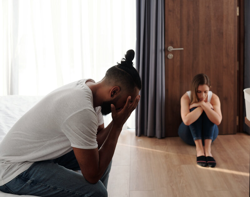 An Interracial couple sitting apart, a male with his head in his hands on a bed and a female sitting on the floor looking regretful and sad – representing relationship support for working through an affair with Freedom Couple Counselling in Melbourne.
