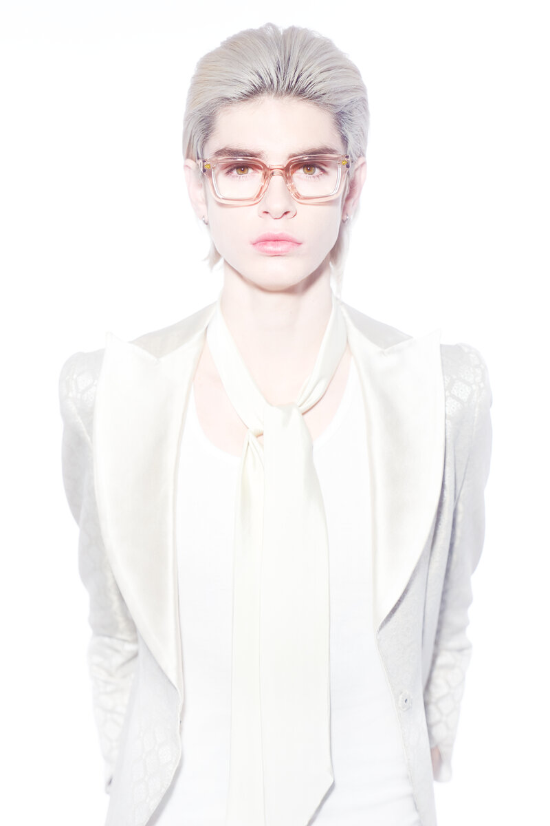 Straight on shot of a blonge woman wearing transparent pink rimmed glasses in front of a plain white background