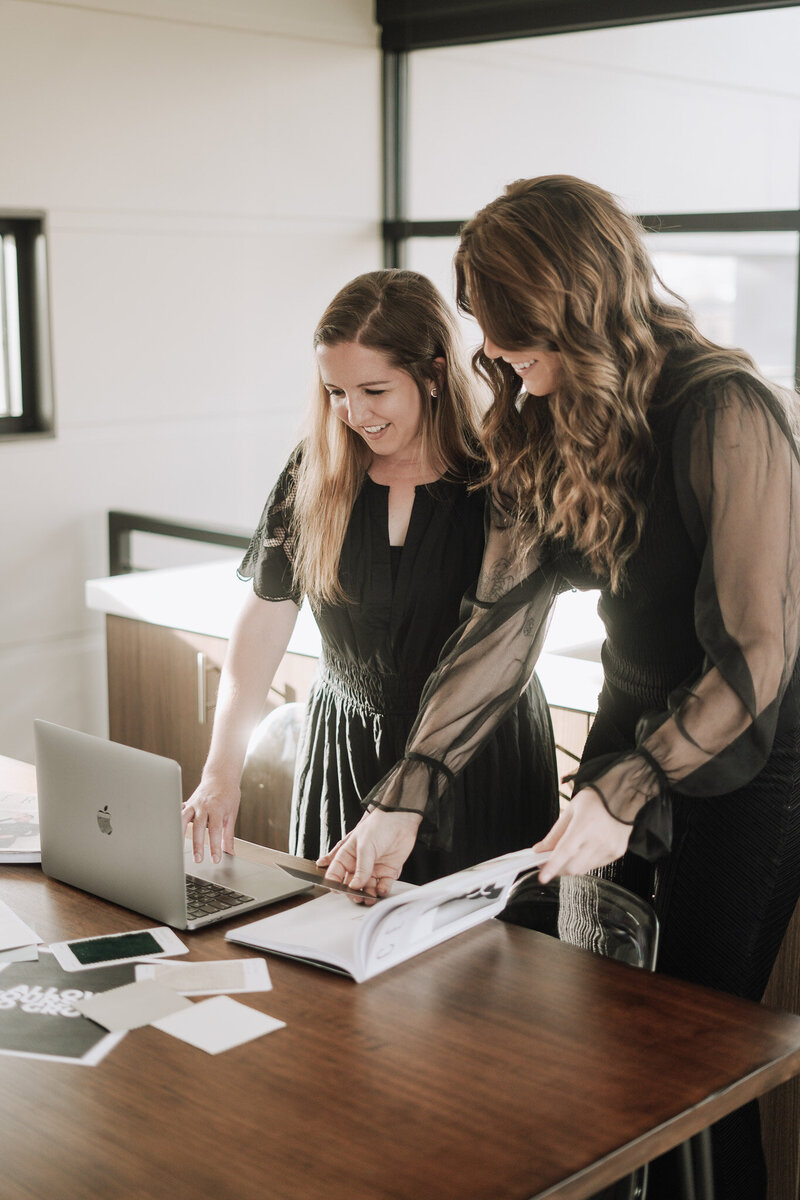 Two women stand at desk with laptop and book as they review branding for a client based in Knoxville