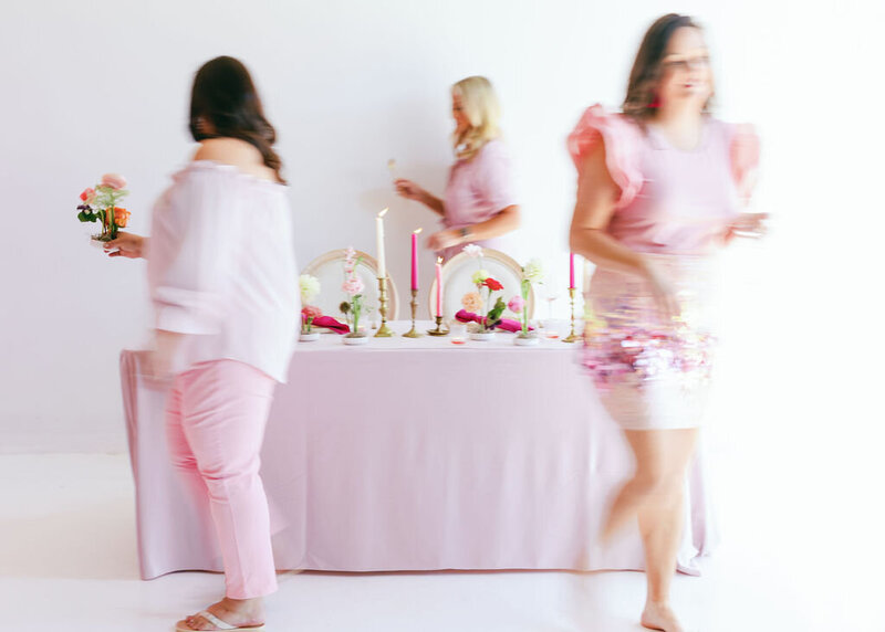 Motion blur of 3 women in pink outfits setting up a table arrangement with pink flowers and candles