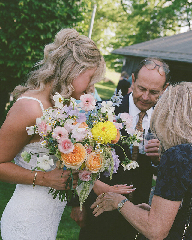A bride in a white dress holds a colorful bouquet of flowers. She interacts with guests outside, creating a joyful and intimate wedding atmosphere.