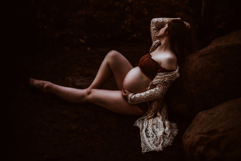 A pregnant woman in a bathing suit and lace cardigan sits in a rock formation illuminated by the setting sun. 