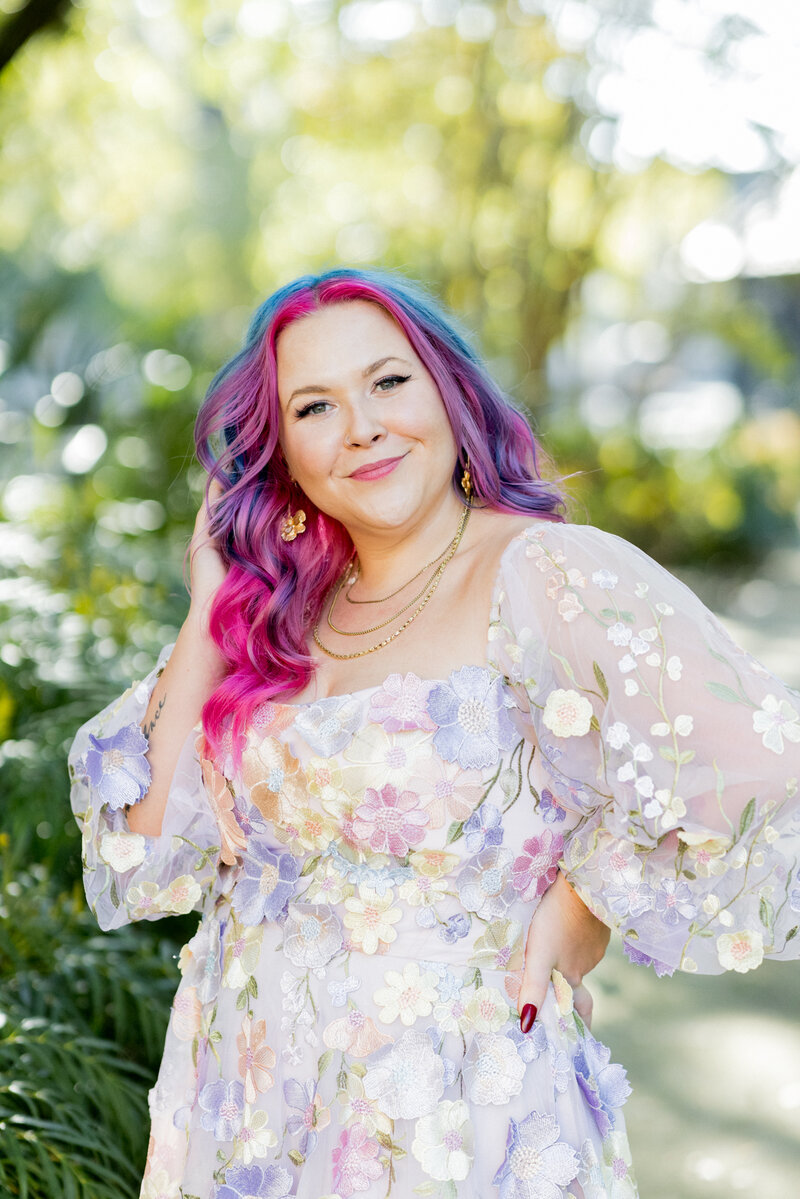 Confident woman with vibrant pink and blue hair poses in a sunlit outdoor setting, wearing a pastel floral embroidered dress with sheer sleeves. Her gold jewelry and bold makeup complement her expressive, creative style. Small business branding photography in Columbia, SC, showcasing unique personal branding for artists, entrepreneurs, and creatives.