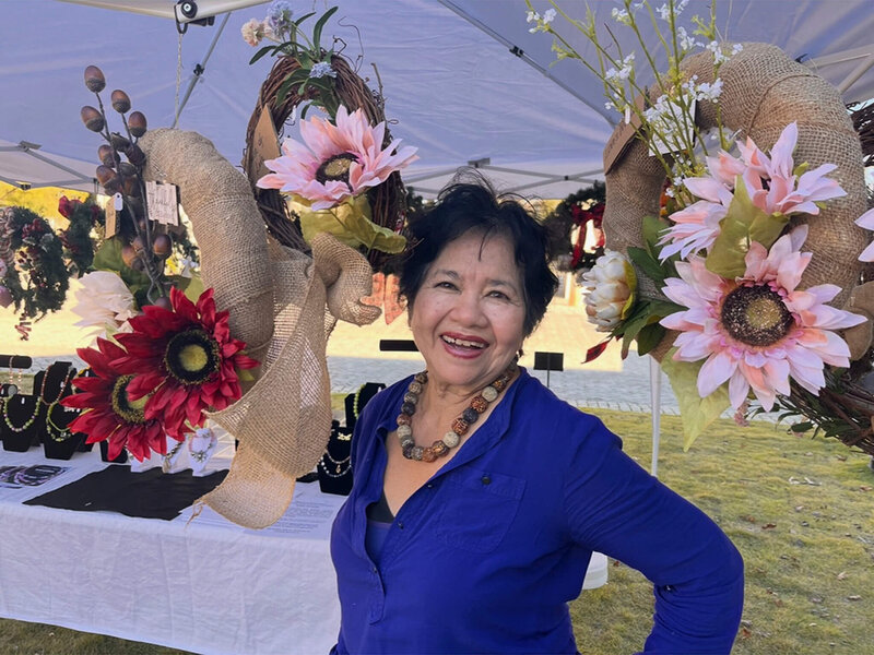 Nita Tin, author, speaker, and life coach from Atlanta, Georgia, smiling under a white tent at a local art fair, standing in front of her Maker's Space handmade burlap wreaths decorated with large pink, red, and white flowers.