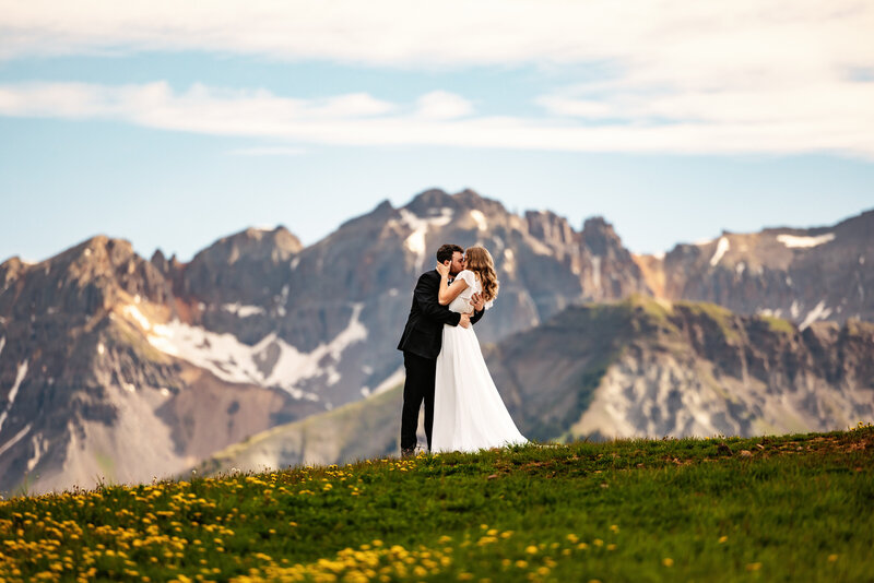 Shannon Lee, a Colorado elopement photographer capturing a romantic kiss between a couple in a stunning mountain landscape with wildflowers in the foreground.