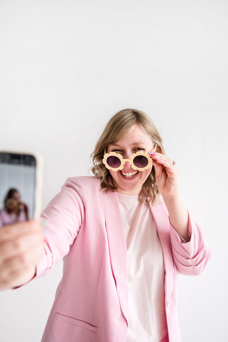 Kat Murphy taking a selfie in a light pink blazer putting on orange flower sunglasses