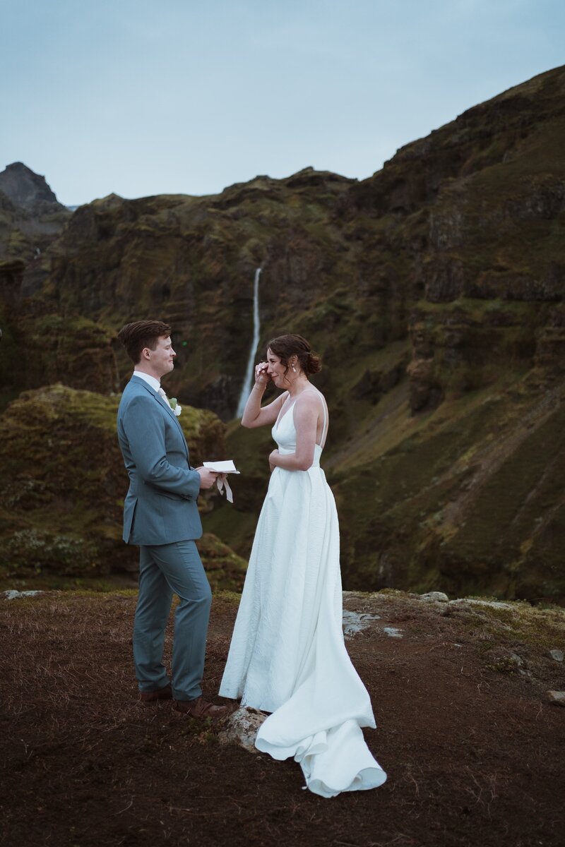 couple is reading vows on a cliff edge overlooking a waterfall. The bride is wiping away a tear, and the groom is holding his vow book. He is in a navy suit and she is in a white dress.