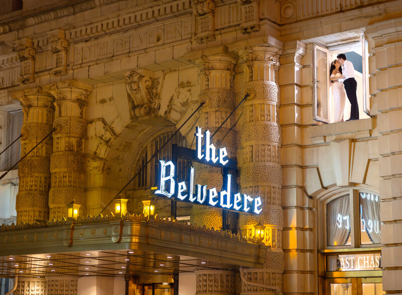A couple stands in the window of the Belvedere Hotel, sharing a romantic kiss. The buildings ornate architecture is illuminated at night, highlighting the hotels name in bright, elegant lettering.