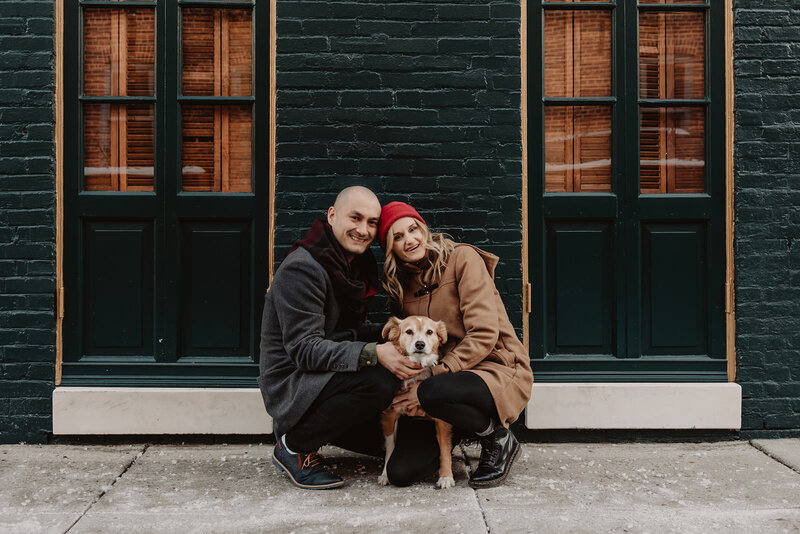 A couple kneeling down, smiling with their dog in between them 