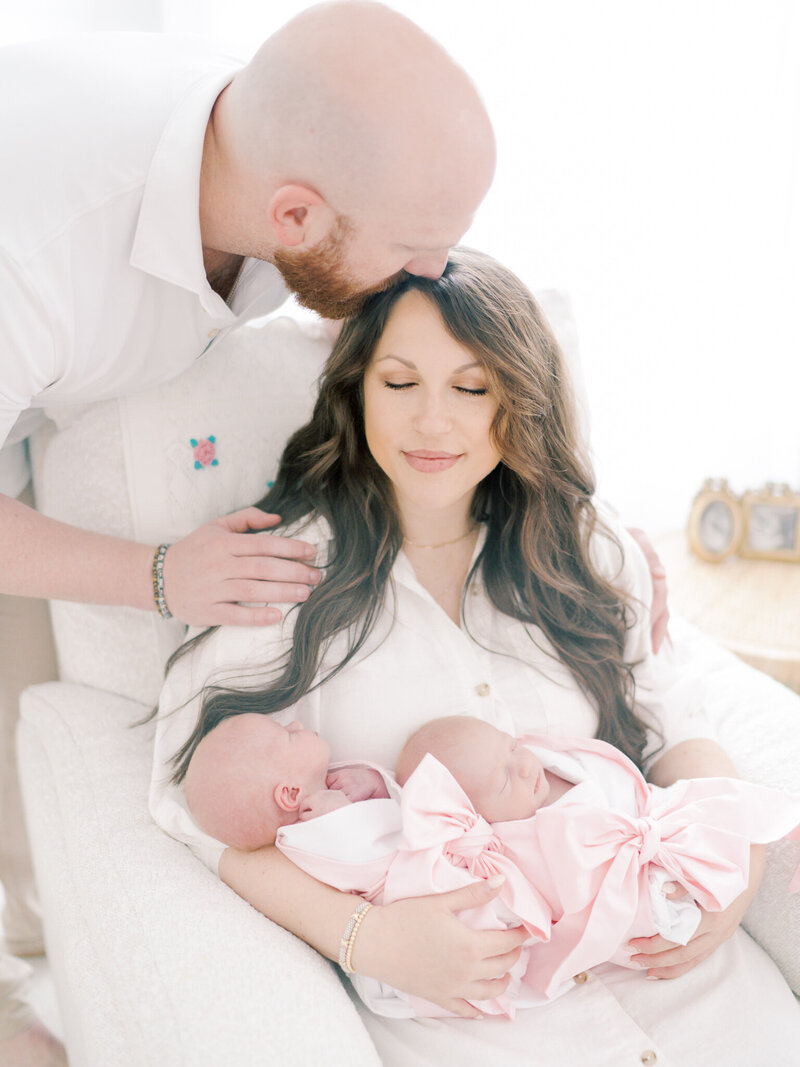 Mother in rocking chair holding twin newborn girls wrapped in swaddles while dad leans down to kiss her head