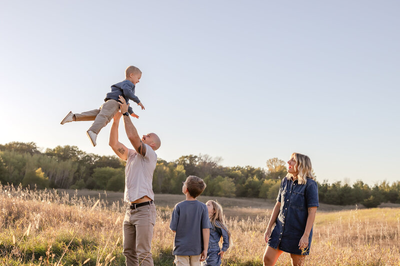 a father throws his young son into the air at a field in Lakeville, Minnesota for a fall family photo shoot. The family of 5 smile at eachother while wearing hues of blue and stand in a grassy field at sunset.