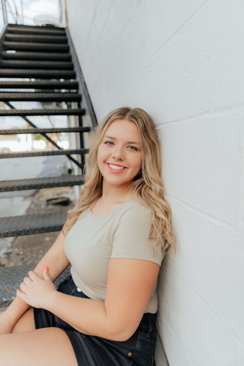 Senior portrait of a smiling high school graduate sitting on outdoor stairs against a white wall, captured in natural light by a Midwest senior photographer serving Michigan and Ohio.