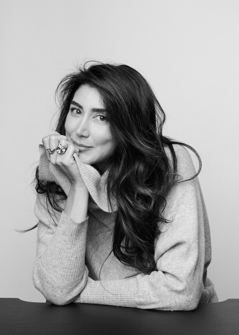 Black and white portrait of a woman in a cozy, oversized sweater, gently resting her chin on her hand with a soft, confident smile. Her long, wavy hair cascades over her shoulder, and she wears a bold statement ring, exuding warmth and elegance. A natural, unfiltered moment from 'THIS IS ME – A Photography Experience by Katie Campbell' in Atlanta, GA, celebrating authenticity and self-expression.