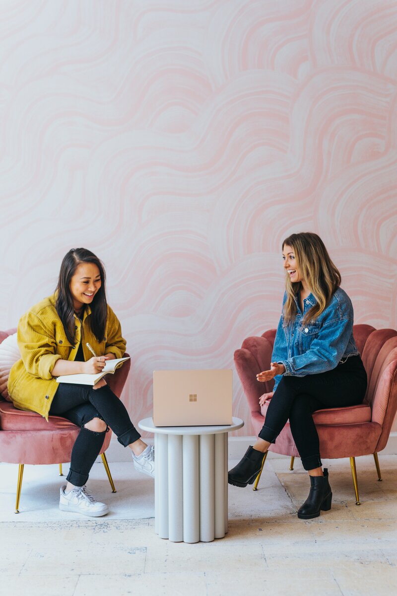 Two women sit in pink chairs with a laptop