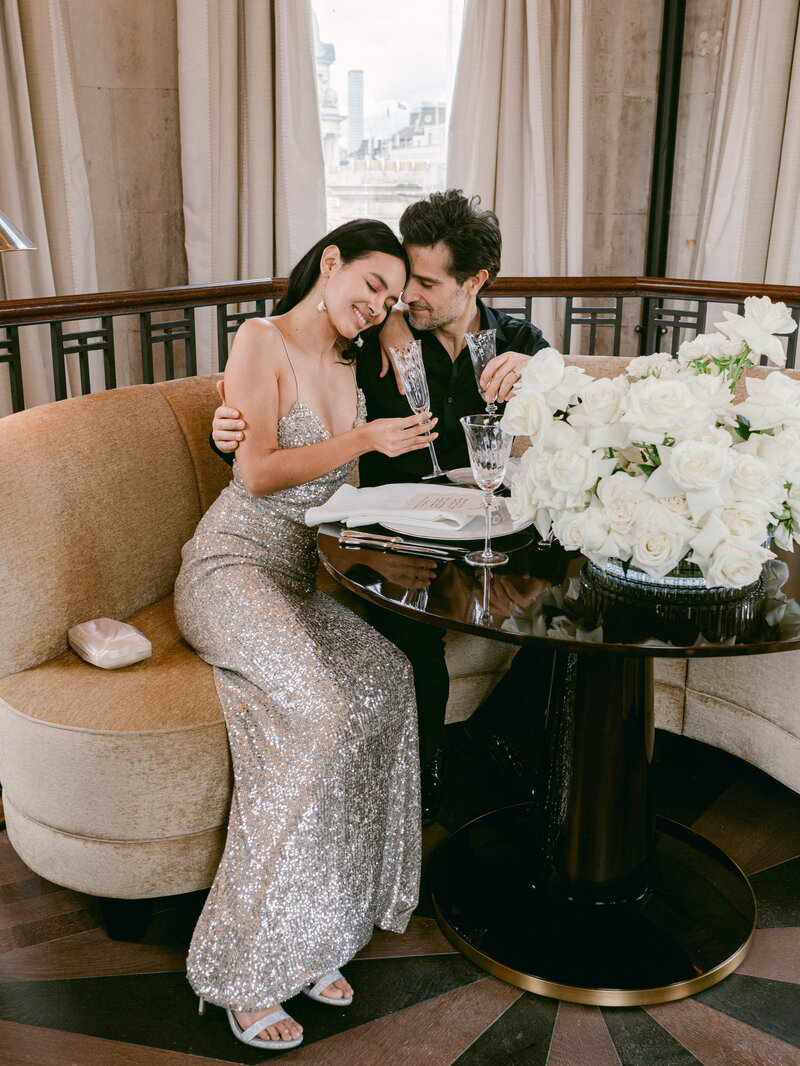 Romantic proposal at Raffles London at the OWO Hotel. The bride-to-be wears a sparkling Monique Lhuillier evening gown, seated with her partner at an elegant table adorned with white roses.