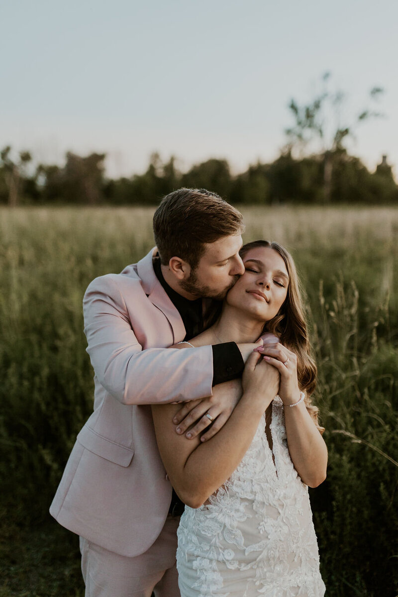 Groom wearing pink suit hugging and kissing bride on cheek from behind
