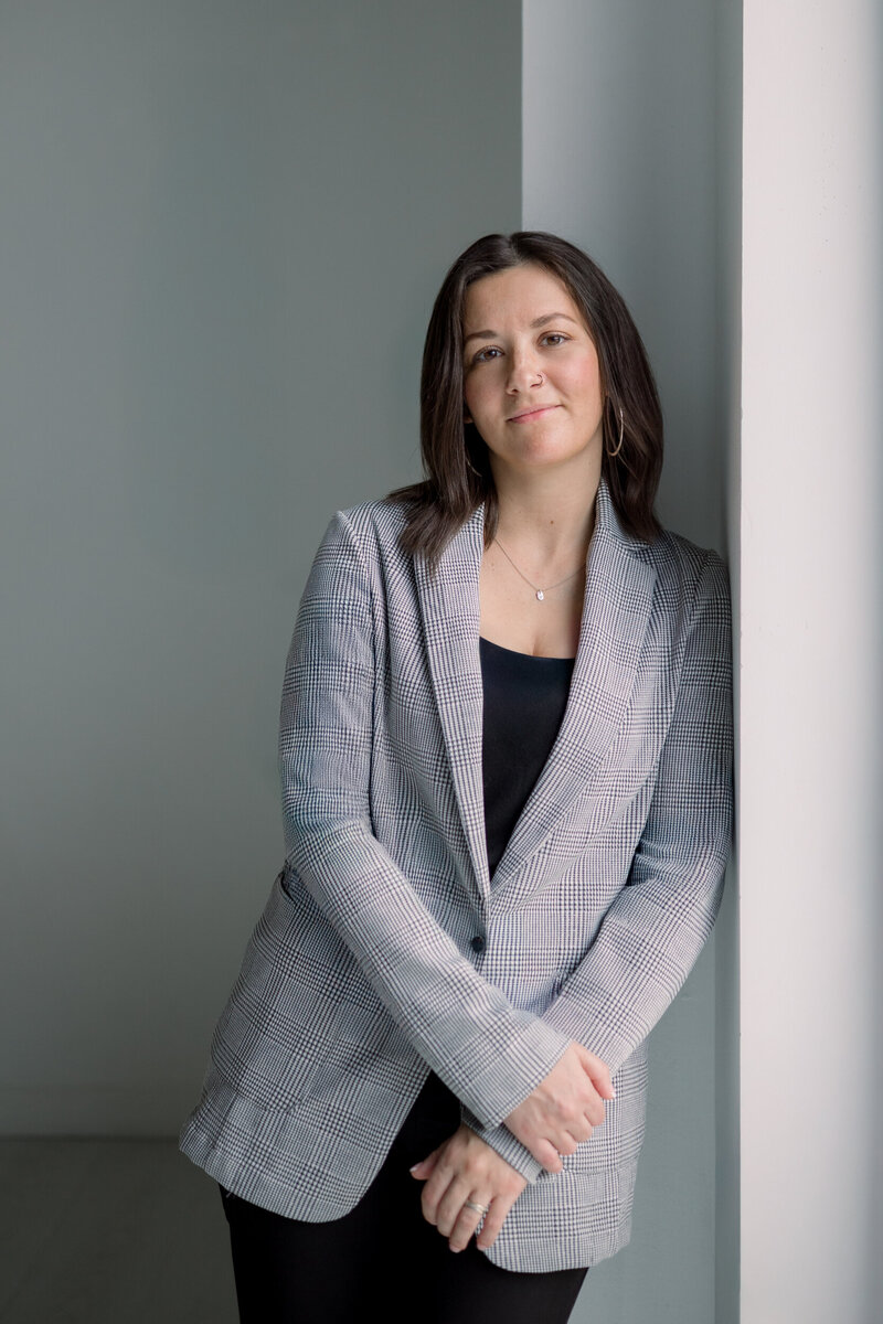 Lois Macomber, San Antonio wedding photographer, standing by a soft gray wall in natural light—capturing her calm, intentional presence and refined, approachable style.