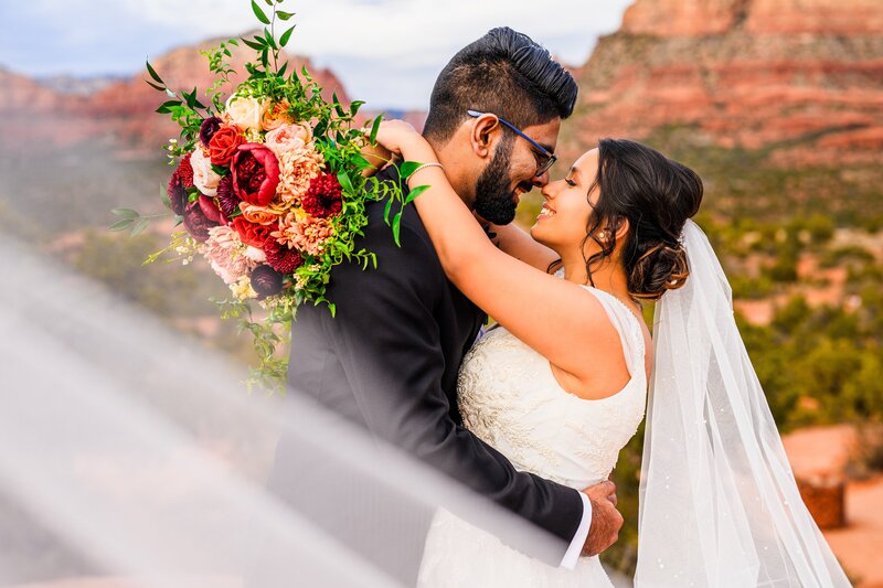 bride and groom touching noses with bouquet over groom's back and veil over lens with red rock background in Sedona Arizona