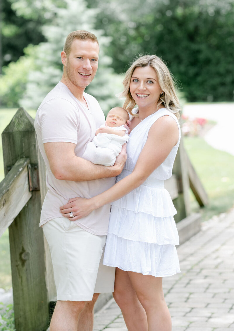 A young couple poses outdoors holding their newborn daughter in a white blanket as they pose for their Evanston  newborn photographer