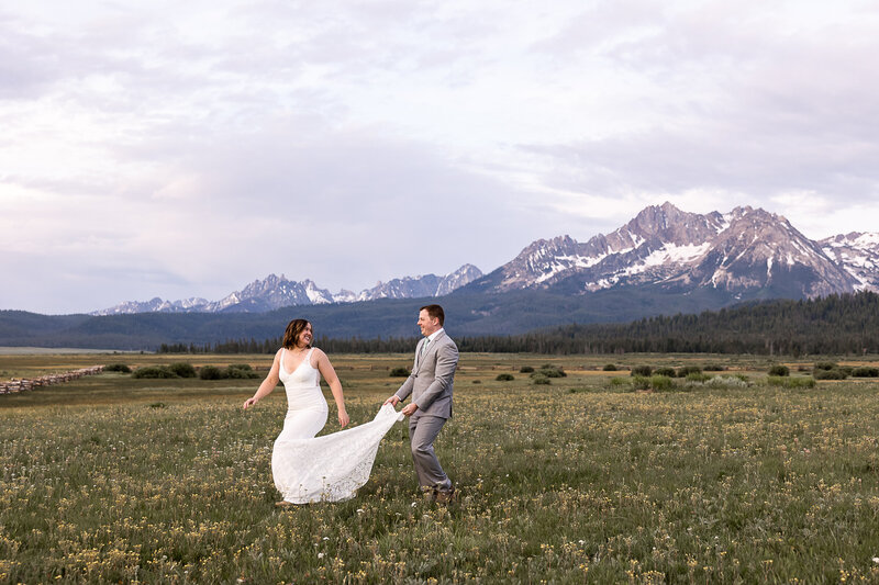 Bride running through a flower meadow with groom running behind holding her dress laughing