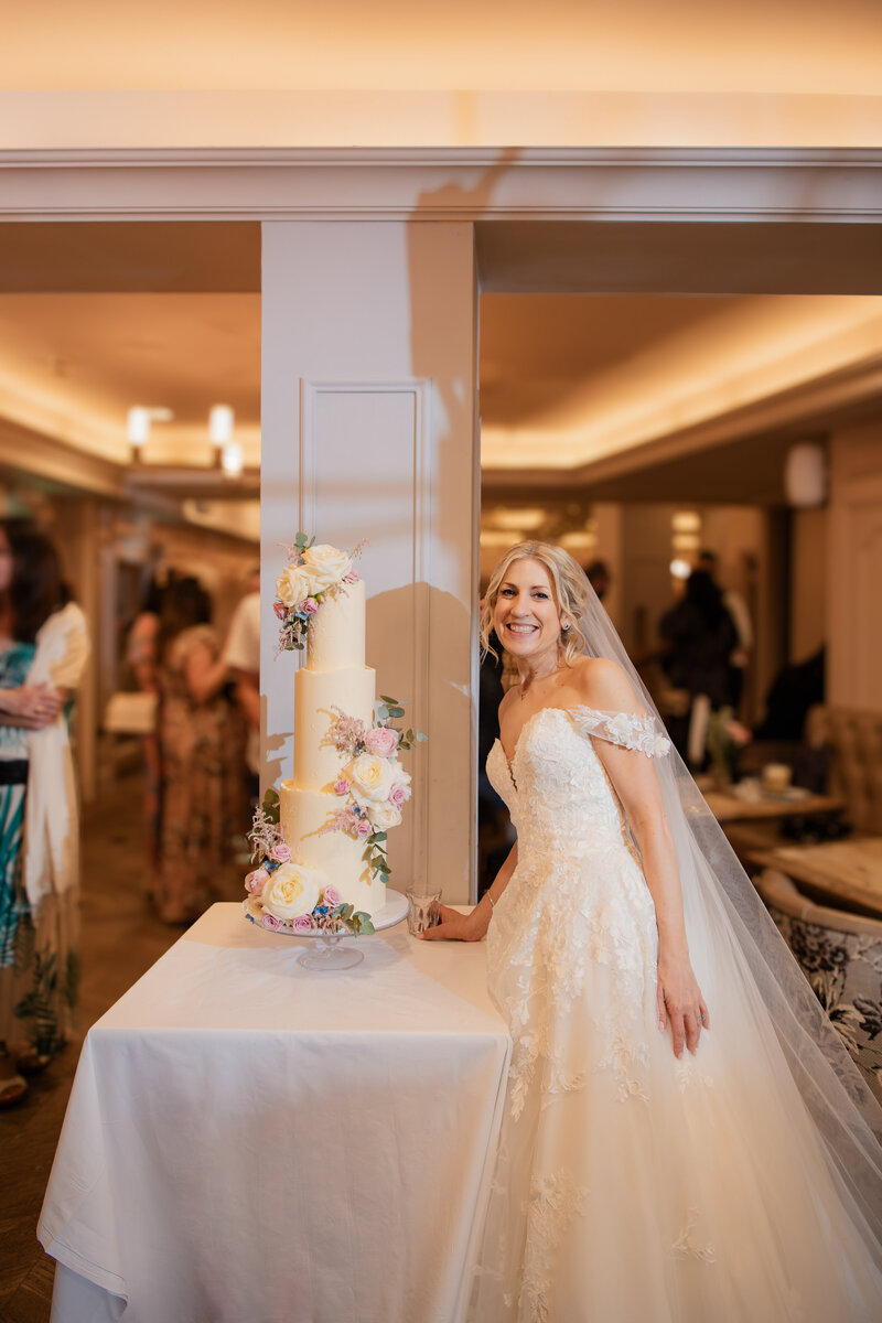 A bride on her wedding day in her wedding dress standing with her three tier wedding cake in  pink and white fresh florals at the Dog & Fox Wimbledon Village