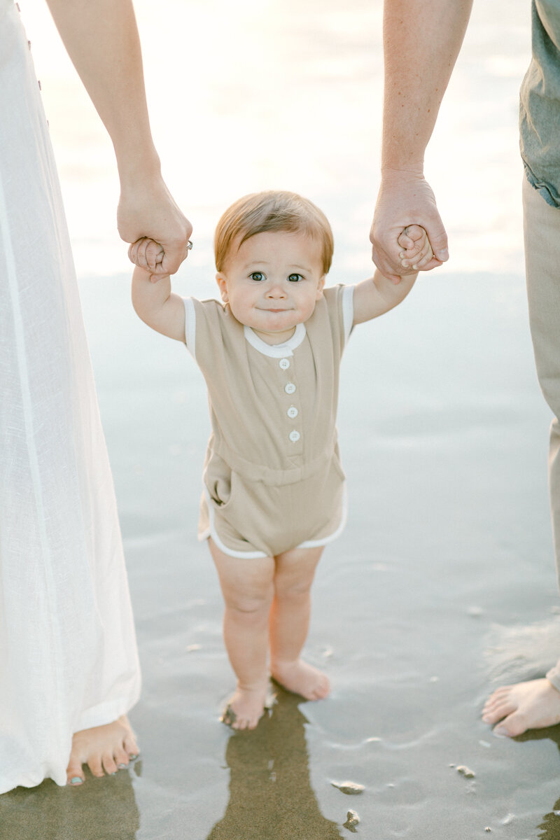 Parents hold toddler's hands as they walk down the beach together during family photography session
