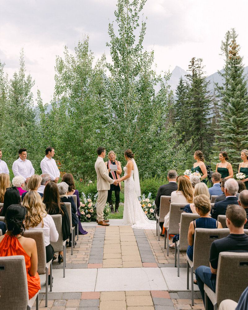 A stunning wedding ceremony at the Malcolm Hotel in Canmore, featuring the bride and groom holding hands as they exchange vows with the iconic Three Sisters mountains in the background.