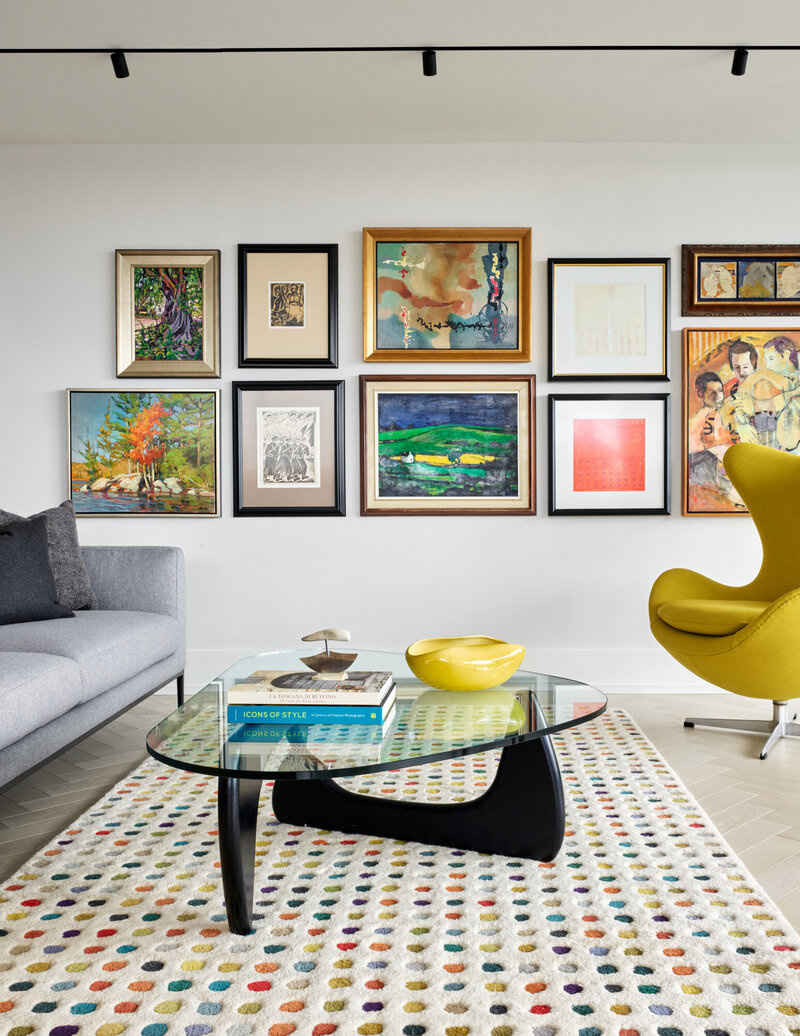 A bright living room featuring a stylish, modern yellow chair, various framed wall-mounted art pieces, a glass coffee table, a soft grey couch, and a colourful patterned rug
