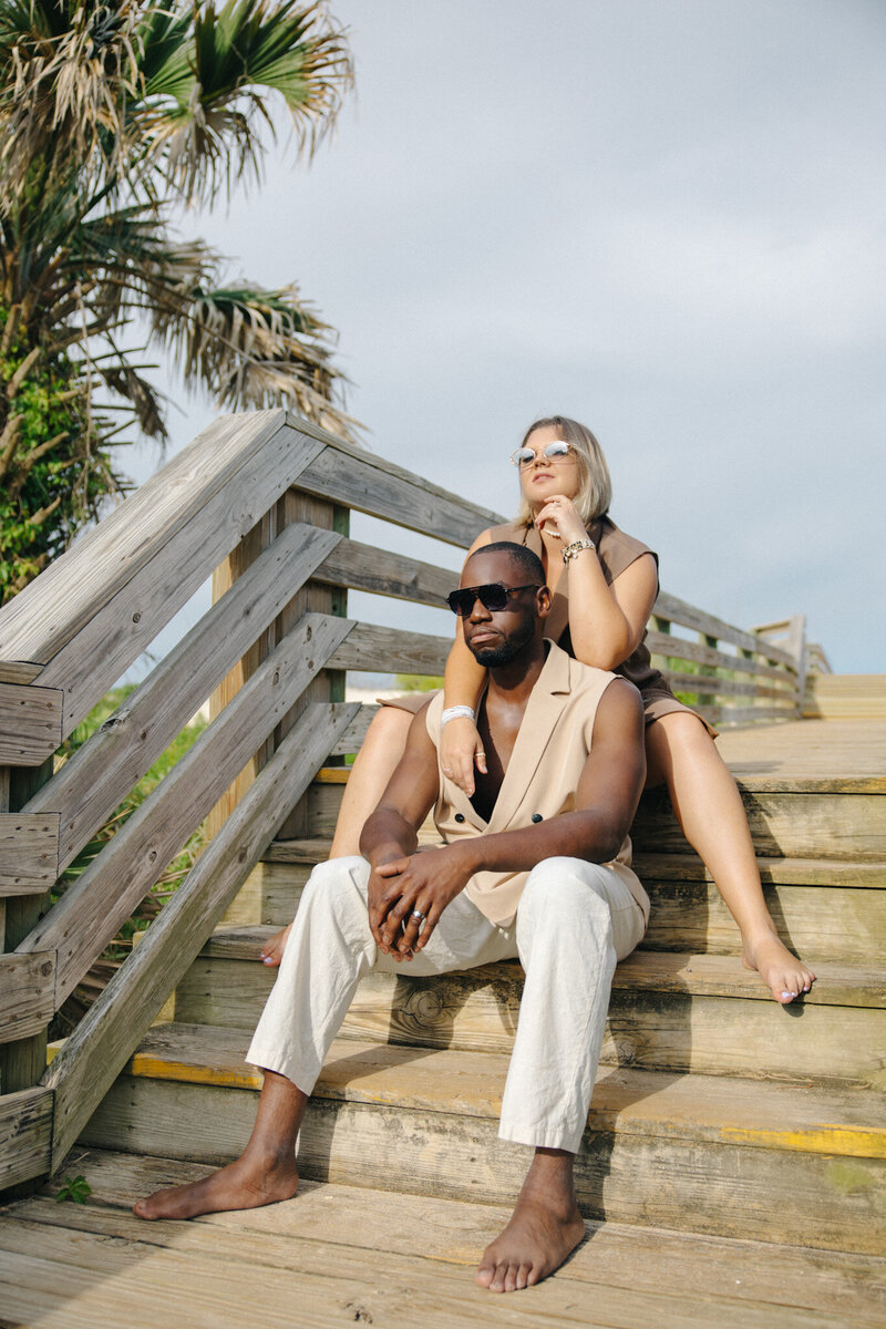 A man and woman wearing sunglasses sitting on wooden steps leading to a beach, framed by palm trees and a blue sky on a sunny day.