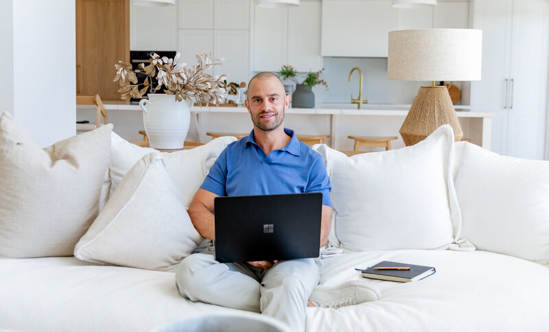 Jon Moses sitting with his laptop, notebook and coffee mug. in front of a white weatherboard wall