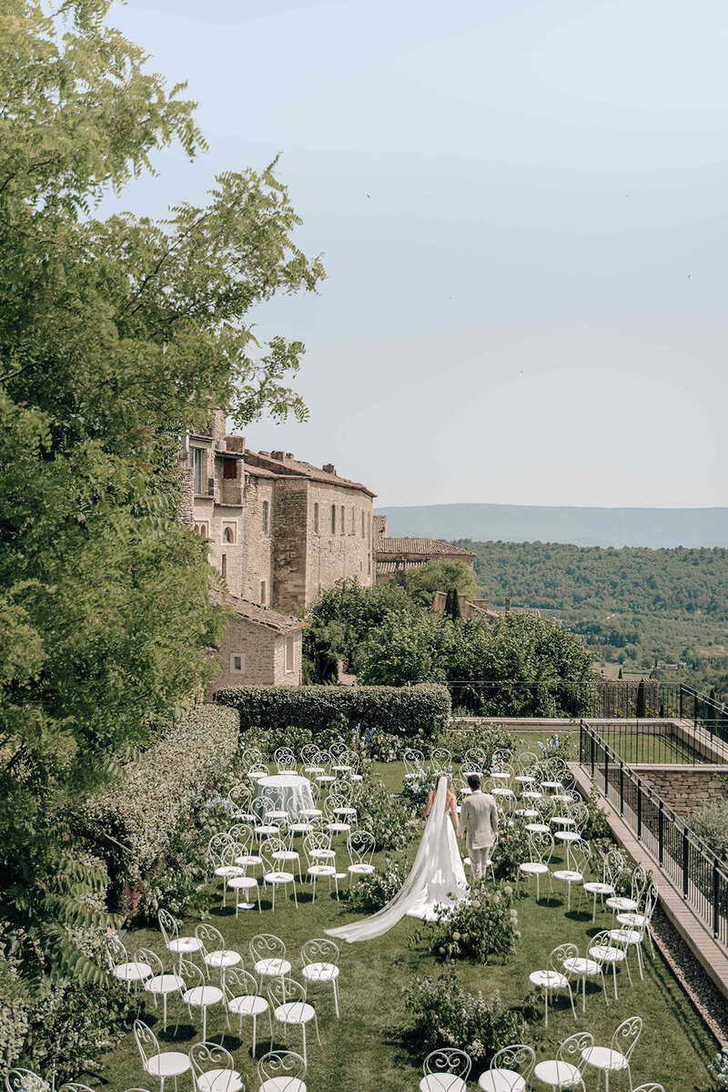 Cliffside terrace  wedding ceremony in a medieval French village with white iron garden chairs in the round, floral arrangements on the ground between chairs and a bride wearing the Lela Rose Avondale Park gown with a cathedral length veil