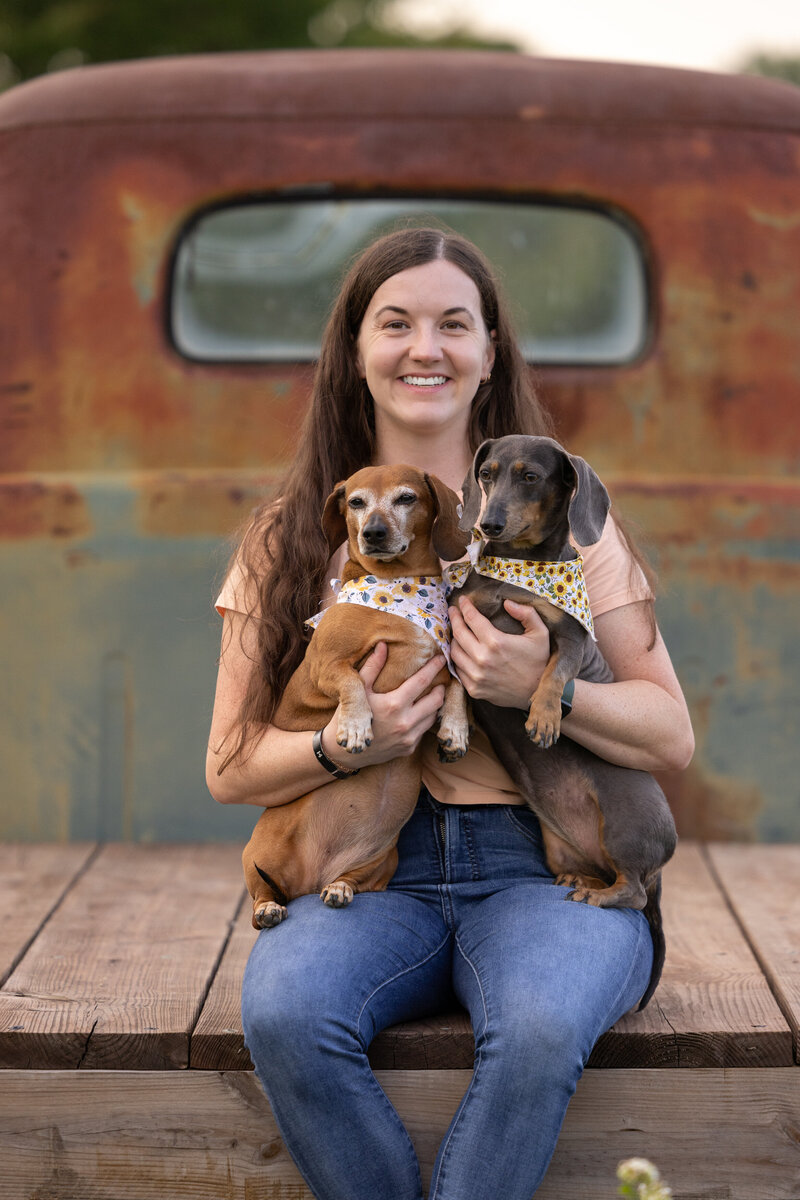A white woman is sitting on the bed of a rusty truck and holding two mini dachshunds. 
