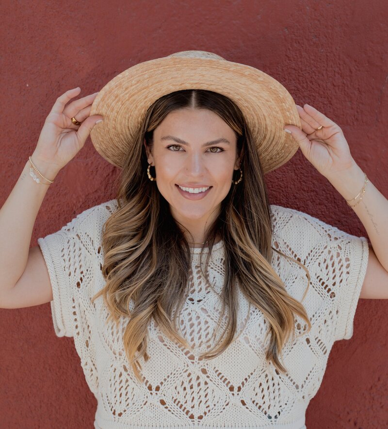 Natalie Bernacchi with long wavy hair wearing a cream knit top and a straw hat, standing in front of a red wall. She holds the brim of her hat with both hands and looks confidently at the camera. The image is styled with a curved corner cutout on the top right. She is sharing her favorite things!