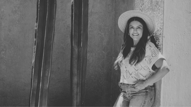 Natalie smiling in a woven top and jeans, leaning confidently against a red wall during a brand photoshoot.
