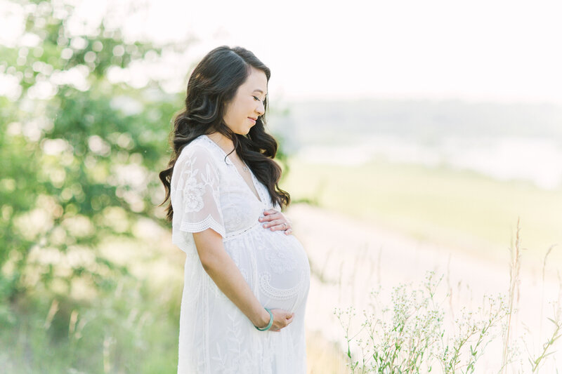 Mother wearing a white dress in a field and holding her baby bump for her Kansas City maternity photos