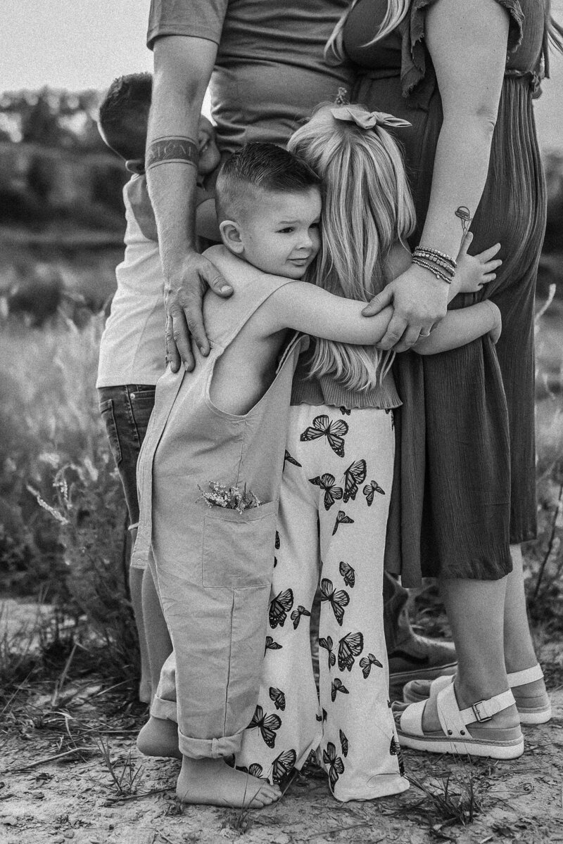 Little boy in overalls with flowers in pockets hugs his family while barefoot during phtoshoot in Olathe KS