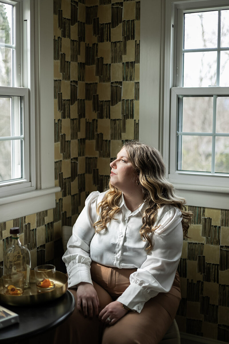 Ashleigh Henry, identity coaching for women leaders, is staring out of a window on a winter's day in Asheville, North Carolina, considering her thoughts in a yellow and green room with old fashioneds by her side