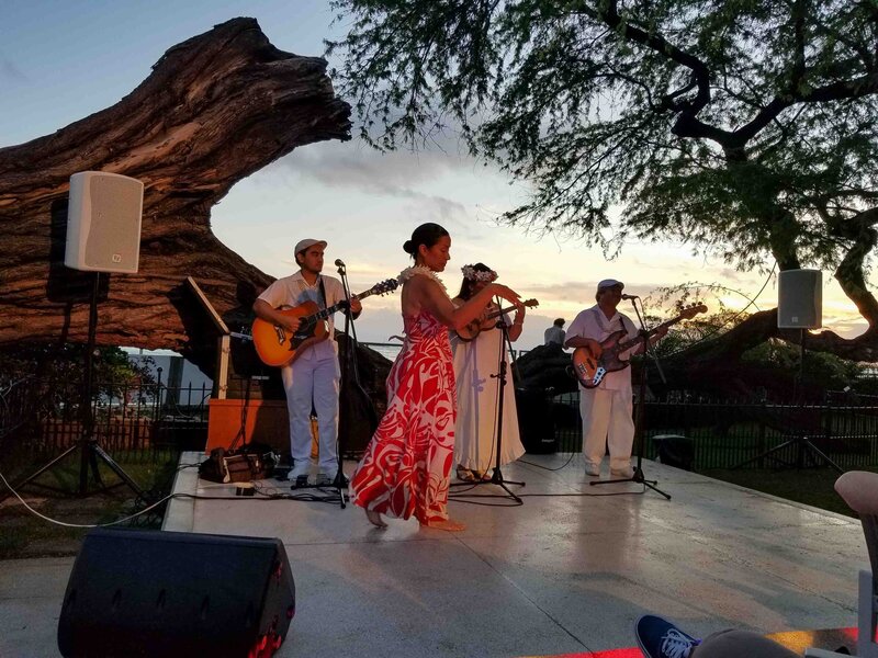 A Hawaiian music group performing at sunset under a sprawling banyan tree in Hawaii, with a hula dancer in a red and white dress and musicians playing guitar and bass ©Stephanie Dosch | theViatrix Curated Hawaii Travel Planning