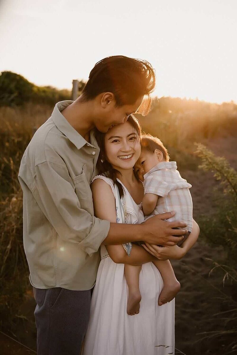 A mother and father holding their young child while the sun sets behind them at a local taranaki beach during their family photoshoot with aimee kelly photography