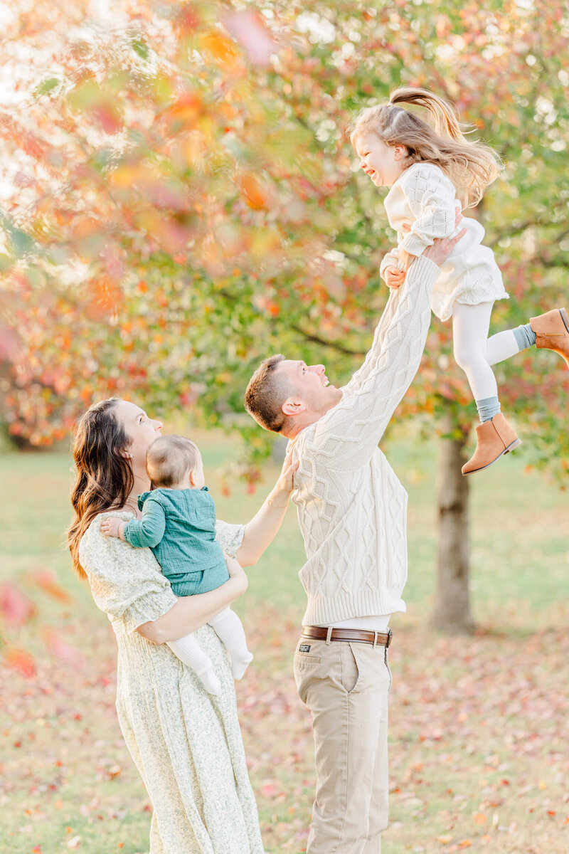 Mom holding baby smiles and looks on as her husband throws their older daughter up into the air