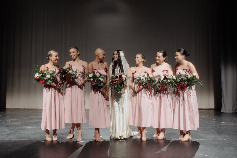 Bride and bridesmaids in pink dresses on stage, smiling together before a theatre wedding ceremony in editorial (solihull) photography style.