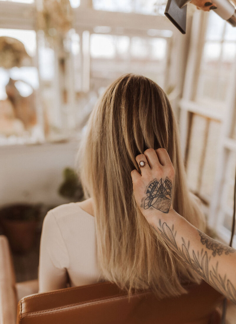 The hair extension expert is styling her client with long straight hair. Both women are in front of a mirror and her hand is tattooed with a butterfly.