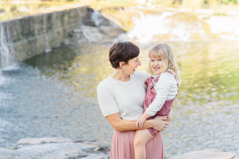 Austin, TX Family & Newborn Photography | A mother and daughter stand in front of a lake at Blue Hole Park. The mother holds her daughter in her arms and they laugh together.