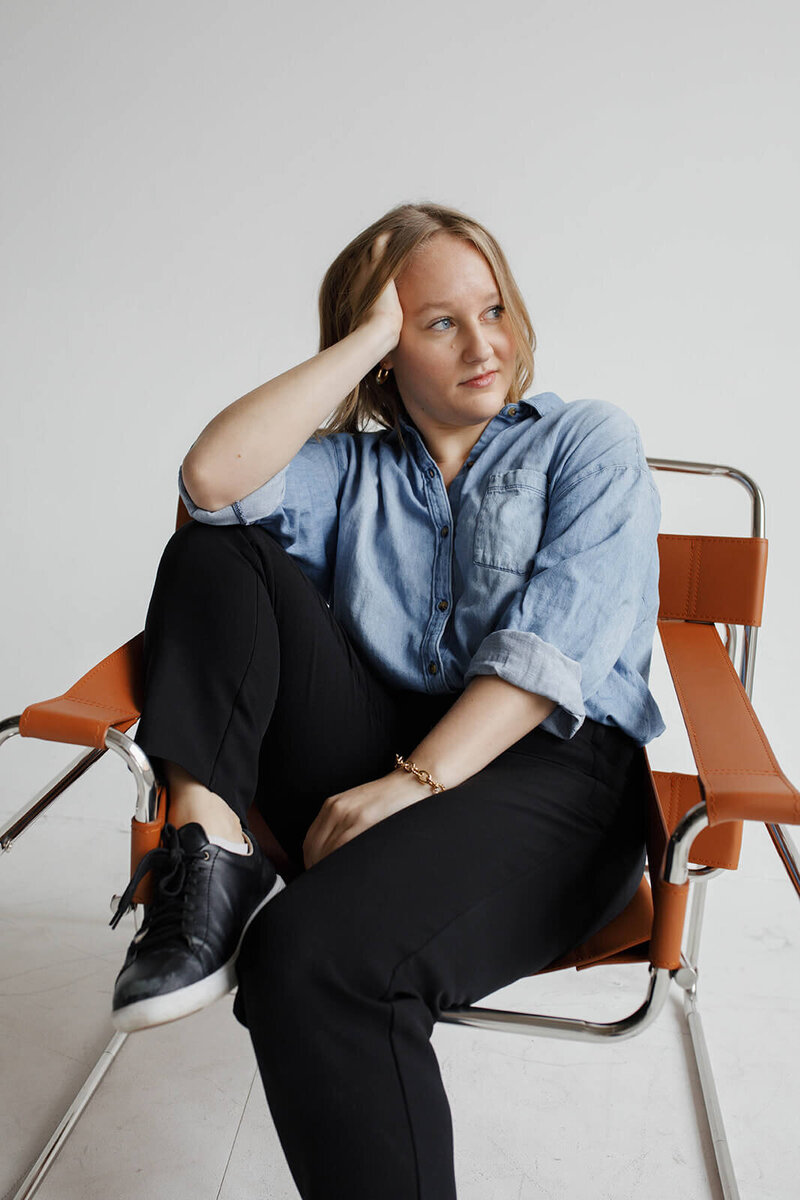 A women in a jean shirt and black pants posing in an orange chair