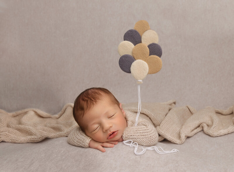 Brooklyn NY newborn photoshoot. Little boy sleeping on his belly with his hands folded under his chin. Under his arm he is holding felt balloons that hare suspended in the air. Captured by premier Brooklyn NY family photographer Chaya Bornstein Photography.