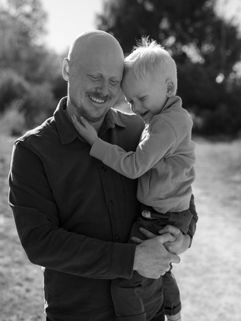 A joyful father shares a playful laugh with his child in this black-and-white candid portrait. The image beautifully captures the deep bond between parent and child, highlighting love and connection through genuine expressions. Photographed by KCP Families