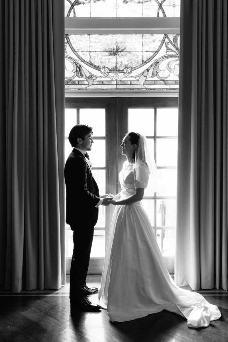 Black and white photo of bride and groom holding hands in front of tall windows, a timeless moment captured at an Austin wedding.