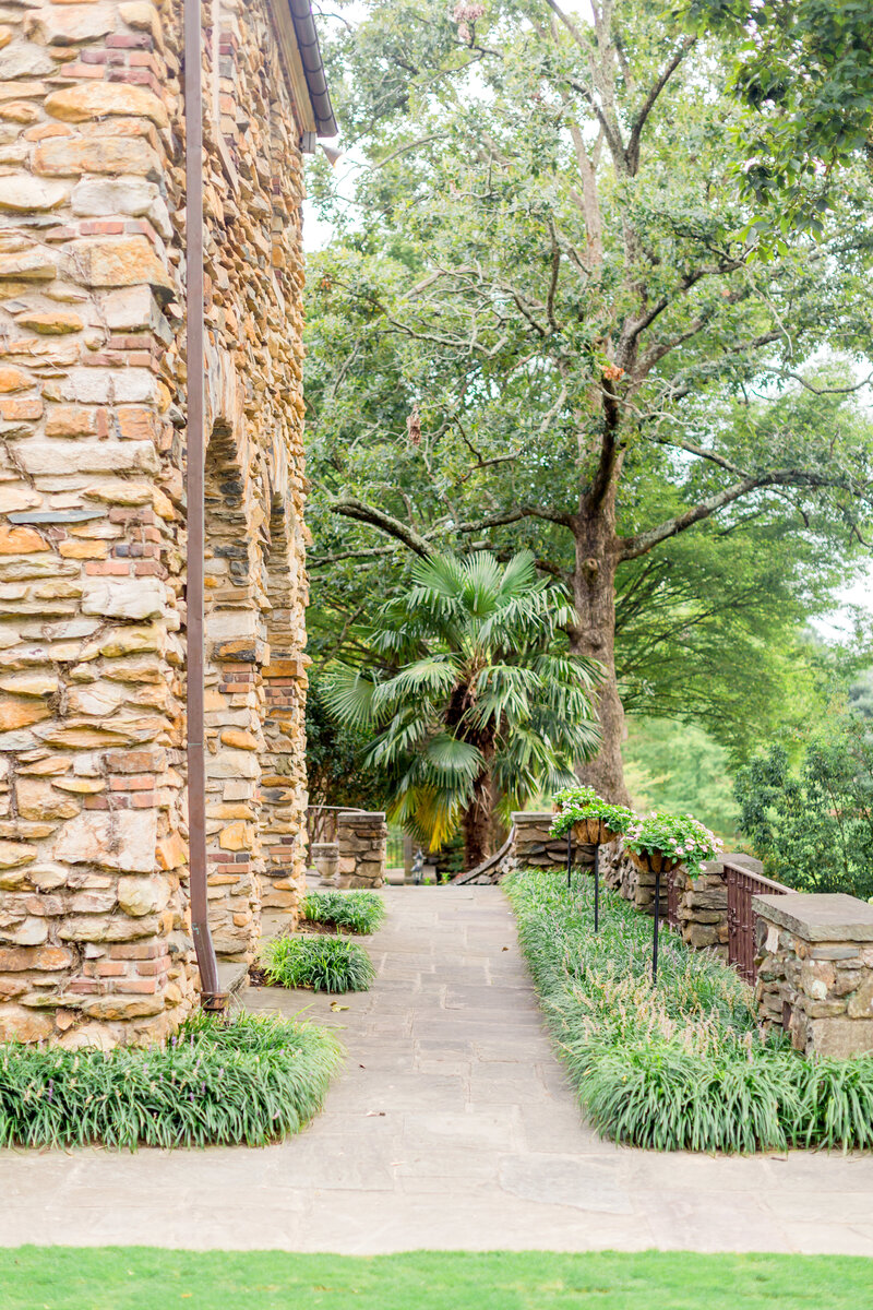 A picturesque stone pathway at Graylyn Estate, lined with lush greenery and blooming flowers, leading to the historic stone manor with timeless old-world charm